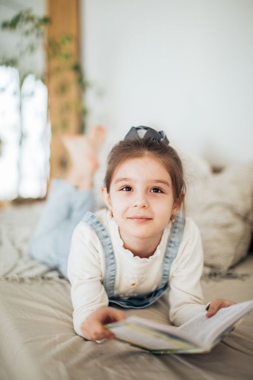 Free A young girl lies on a bed reading a book, creating a cozy indoor scene. Stock Photo