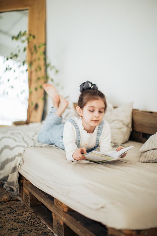 Free Young girl reading a book indoors on a cozy bed. Bright and relaxed atmosphere. Stock Photo