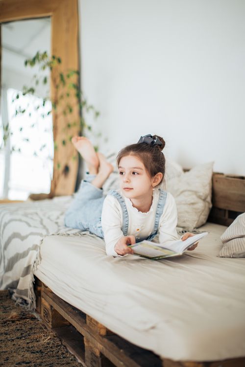 Free A young girl in a cozy room enjoying reading a book while lying on a bed. Stock Photo