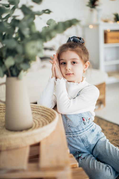 Free Cute child in denim overalls sitting indoors, looking at camera with a warm, artistic background. Stock Photo