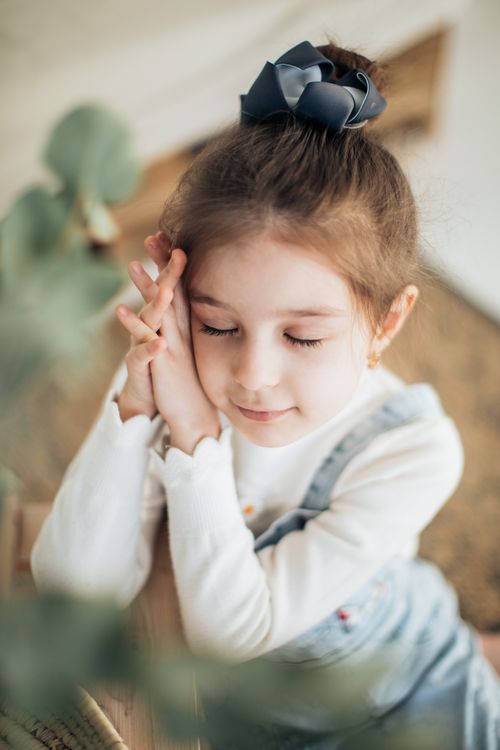Free Charming child in a white sweater and denim overalls, eyes closed, hands together in a serene moment. Stock Photo