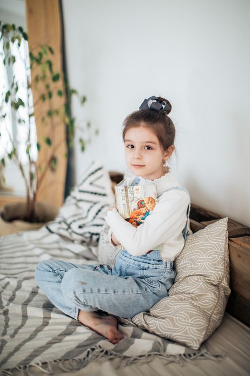 Free A young girl in casual clothing sits comfortably indoors holding a book, showcasing a cozy and peaceful setting. Stock Photo