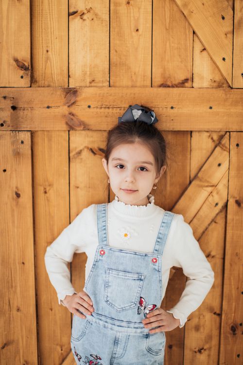 Free Young girl standing with hands on waist against a wooden wall wearing a denim jumper. Stock Photo