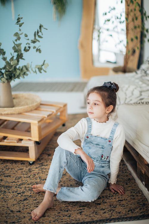Free A young girl in a white jumper and blue jeans sits on a carpet in a cozy, well-decorated room. Stock Photo