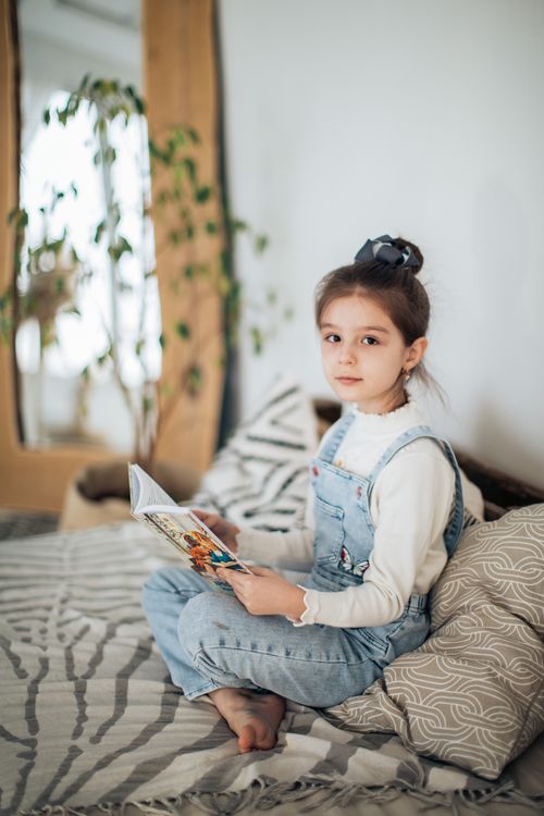 Free A young girl sits comfortably indoors reading a book, surrounded by cozy decor and soft lighting. Stock Photo
