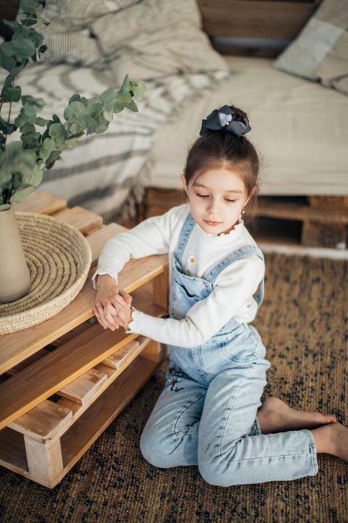 Free Young girl in overalls sitting on carpeted floor, surrounded by rustic wooden decor. Stock Photo