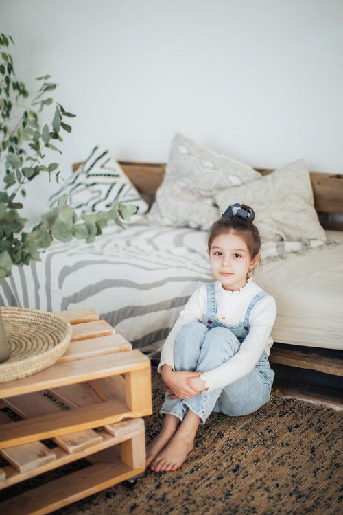 Free A young girl in overalls sitting comfortably in a cozy living room with warm decor. Stock Photo