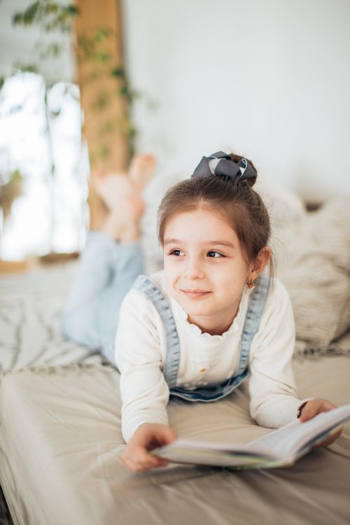 Free Adorable girl with hair bun smiles lying on bed while holding a book indoors. Stock Photo