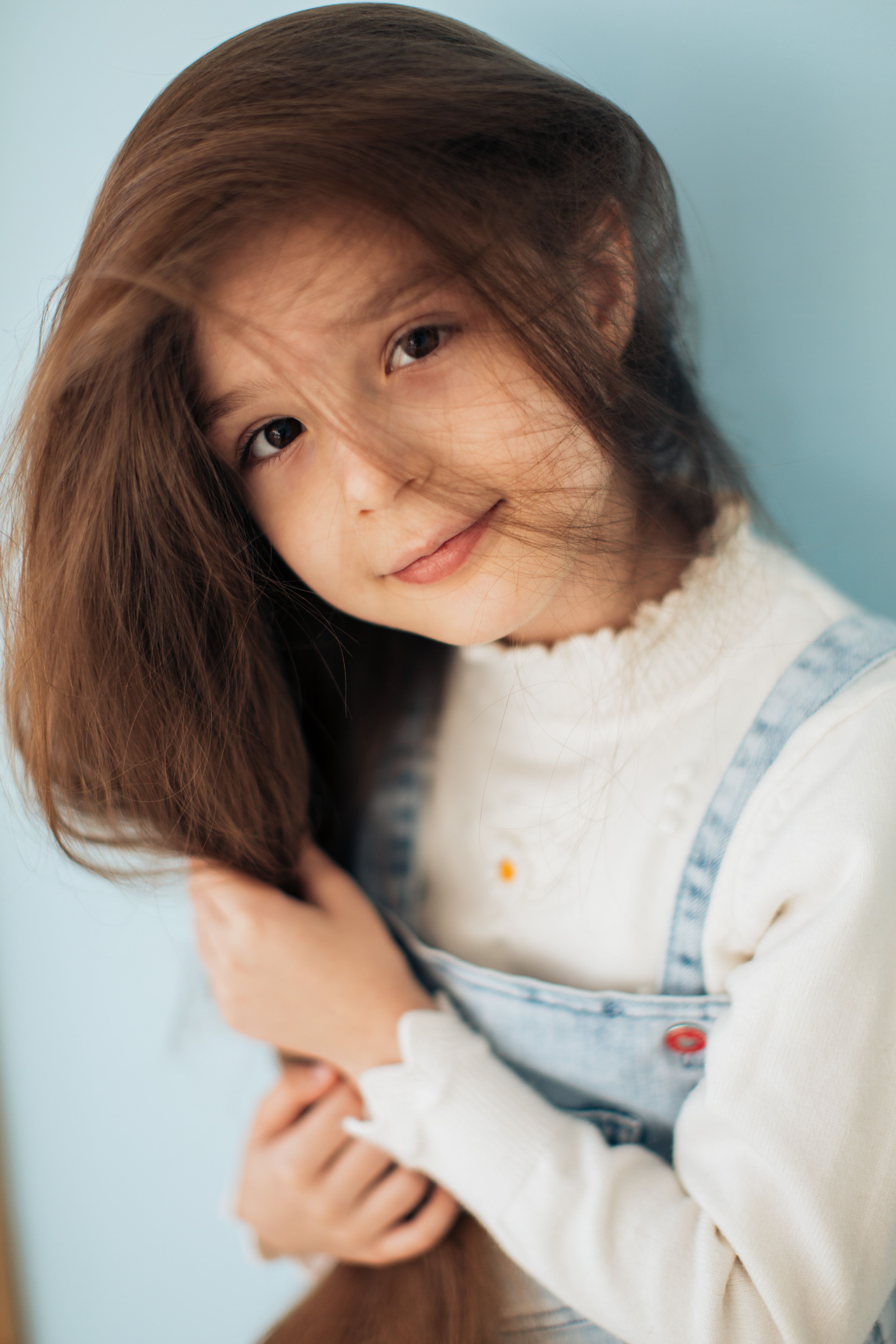 Charming young girl with messy hair wearing denim overalls and white sleeves, smiling softly.