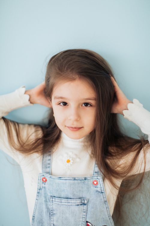 Free Charming portrait of a young girl with long hair in denim overalls against a blue background. Stock Photo