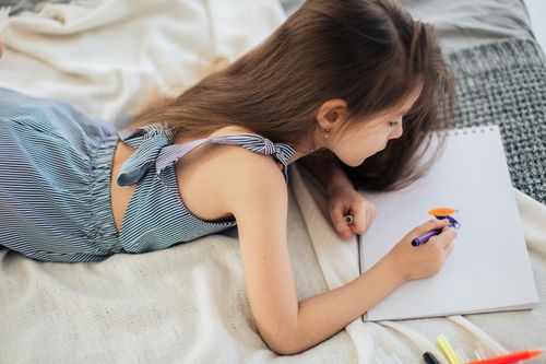 Free A young girl in a striped dress lying down, drawing with markers on white paper. Stock Photo