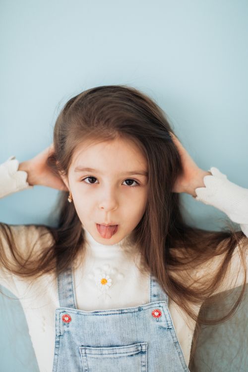 Free A young girl with long hair playfully sticks out her tongue, wearing denim overalls, set against a soft blue background. Stock Photo
