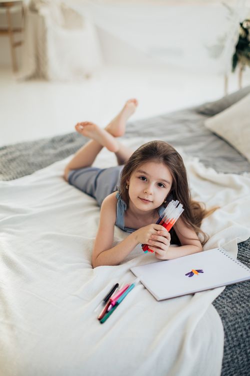 Free A child happily coloring with markers on a bed in a bright and cozy bedroom. Stock Photo