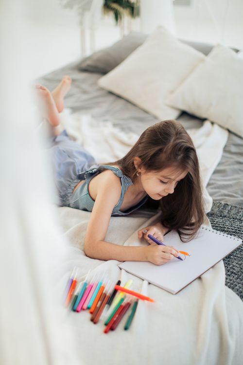 Free Young girl lying on bed, drawing with colored pencils in a cozy bedroom setting. Stock Photo