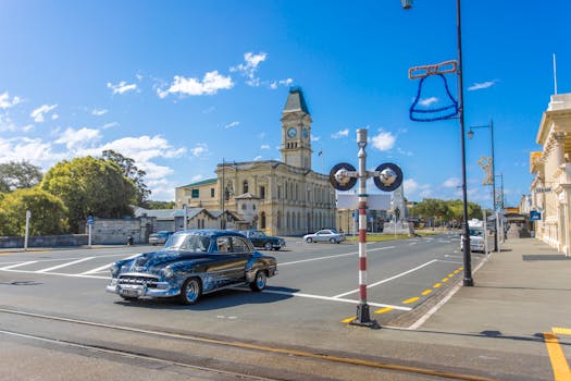 Classic car cruising past historic architecture in Oamaru, New Zealand with clear blue skies.
