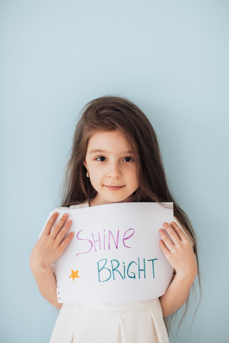 Little Girl Holding A Piece Of Paper With Shine Bright Written On It