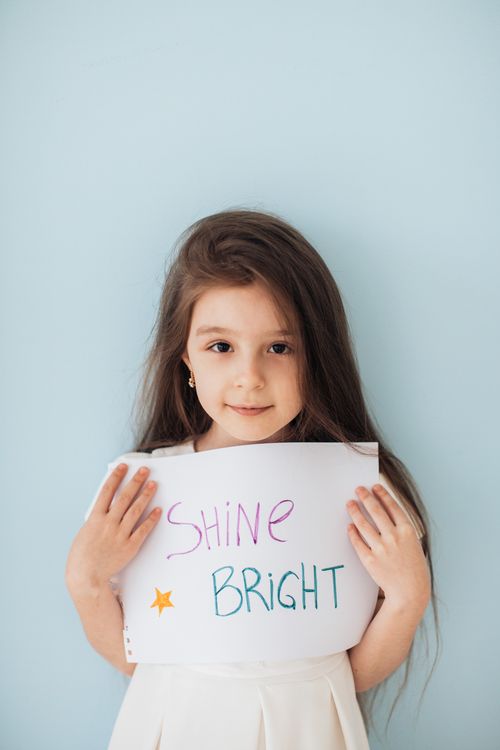Free Young girl holding a motivational sign reading 'Shine Bright' against a light blue background. Stock Photo