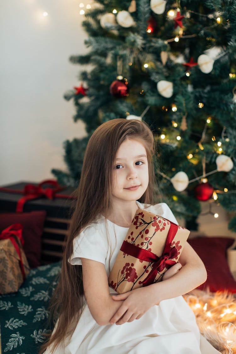 Girl With Long Hair Wearing White Dress Holding A Christmas Present, Decorated Christmas Tree In Background