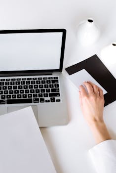 Overhead view of a minimalist workspace with a laptop, tablet, and vases on a white desk.