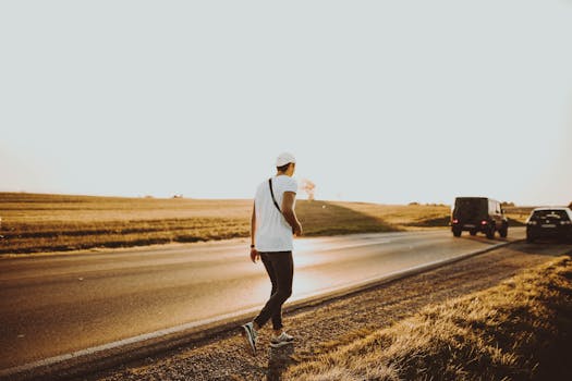 A person walking along a rural road at sunset, with vehicles in the distance under a clear sky.