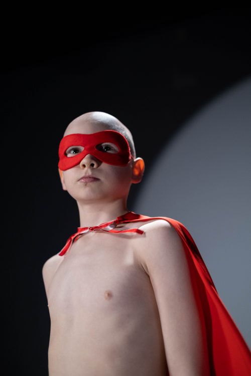 Free A bald child confidently poses as a superhero wearing a red mask and cape in a studio. Stock Photo