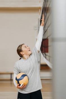 A young female athlete smiling while reaching for a volleyball net indoors.