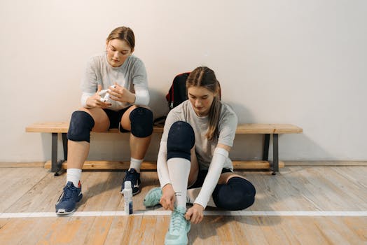 Two female volleyball players in sports gear prepare for a game, sitting on a wooden bench.