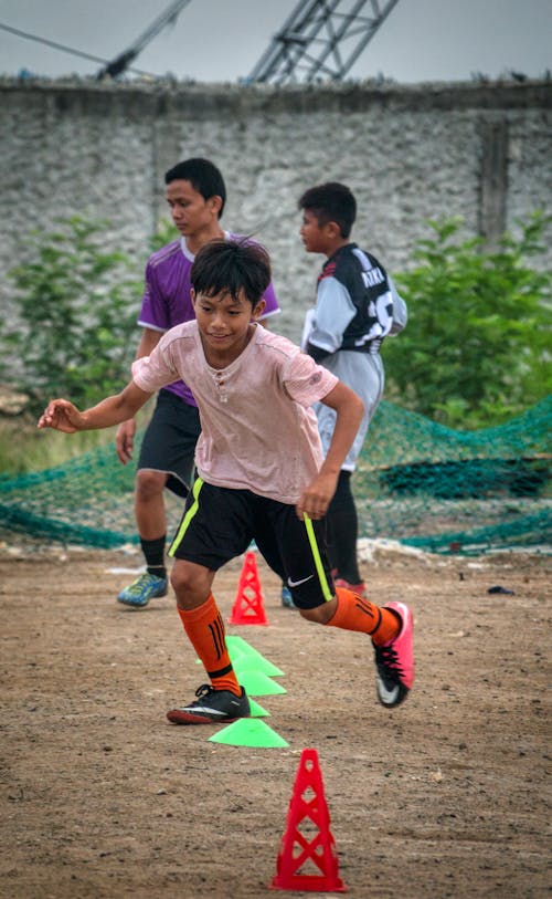 Ethnic kid running through training cones while playing football with team players on field
