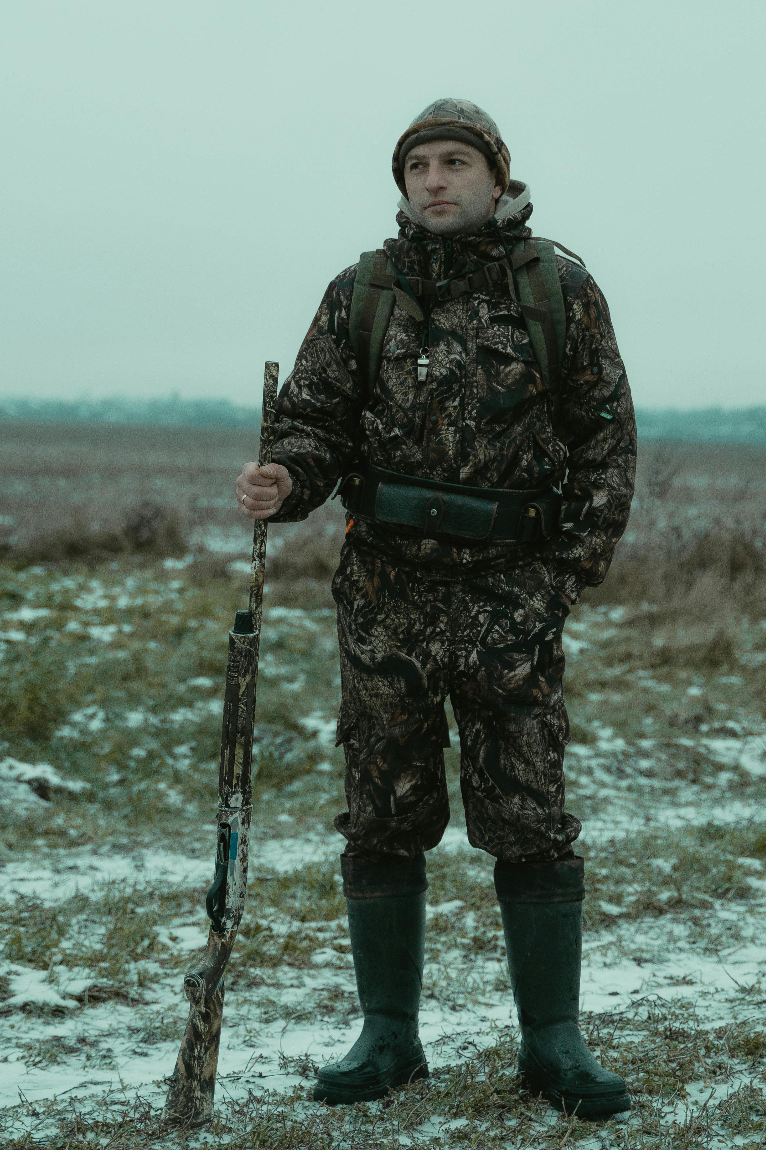 Man in camouflage holding a rifle in a snowy field, showcasing hunting attire and preparedness.