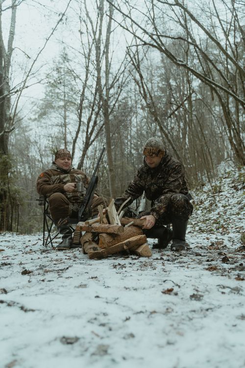 Free Two hunters in camouflage gear set up a campfire in a snowy winter forest environment. Stock Photo