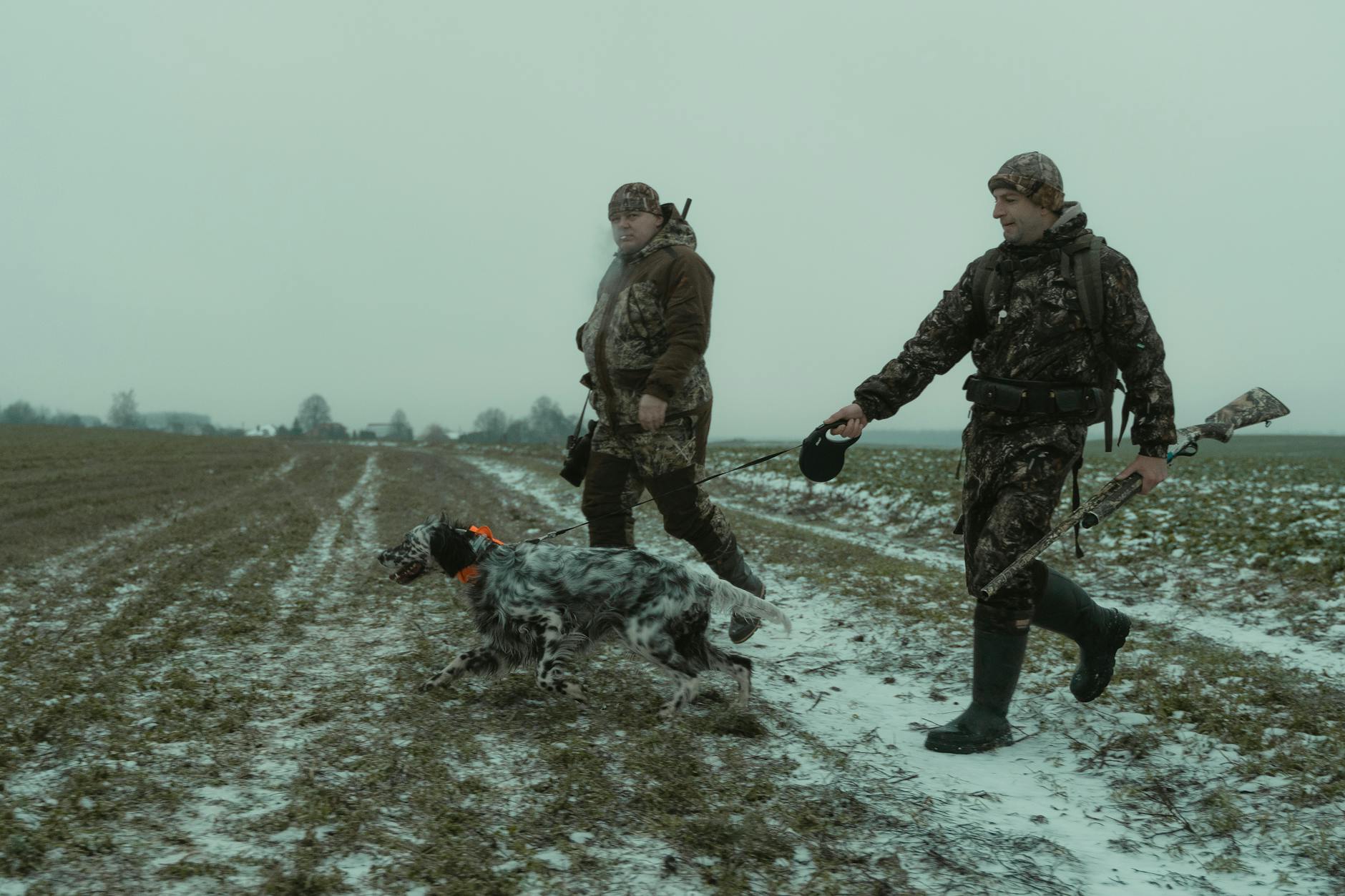 Hunters in camouflage walk with a dog in a snowy field, showcasing outdoor winter hunting expedition.