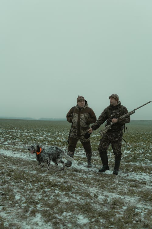 Two Men in Black and Gray Camouflage Uniform Holding Black and White Dog on Gray Field