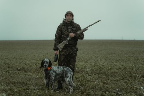 Free A hunter in camouflage gear stands with a dog in an expansive field, showcasing outdoor adventure. Stock Photo