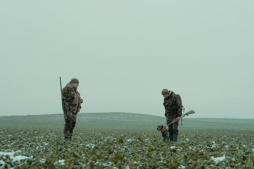 Two hunters with rifles and a dog in a snowy countryside field.