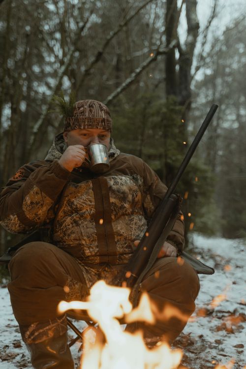 Free Hunter in camouflage enjoys a warm drink by the fire in a winter forest. Stock Photo