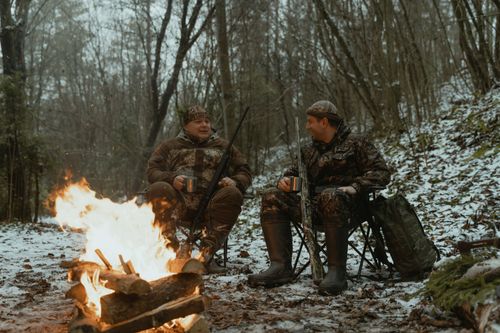Free Two hunters in camouflage sit by a campfire in a snowy forest, sipping hot drinks. Stock Photo