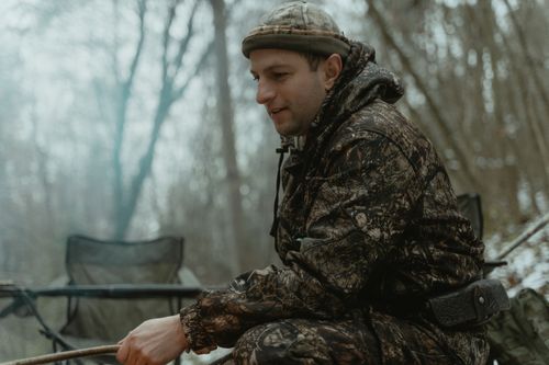 Free A man in camouflage gear sits in a serene forest, blending seamlessly with nature. Stock Photo