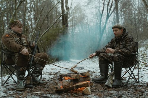 Free Two hunters in camouflage sit by a snowy campfire, roasting sausages in the winter forest. Stock Photo