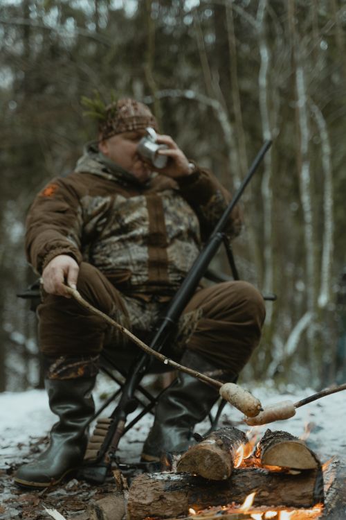 Free Man in camouflage enjoys campfire snack and drink during winter hunting trip in the forest. Stock Photo