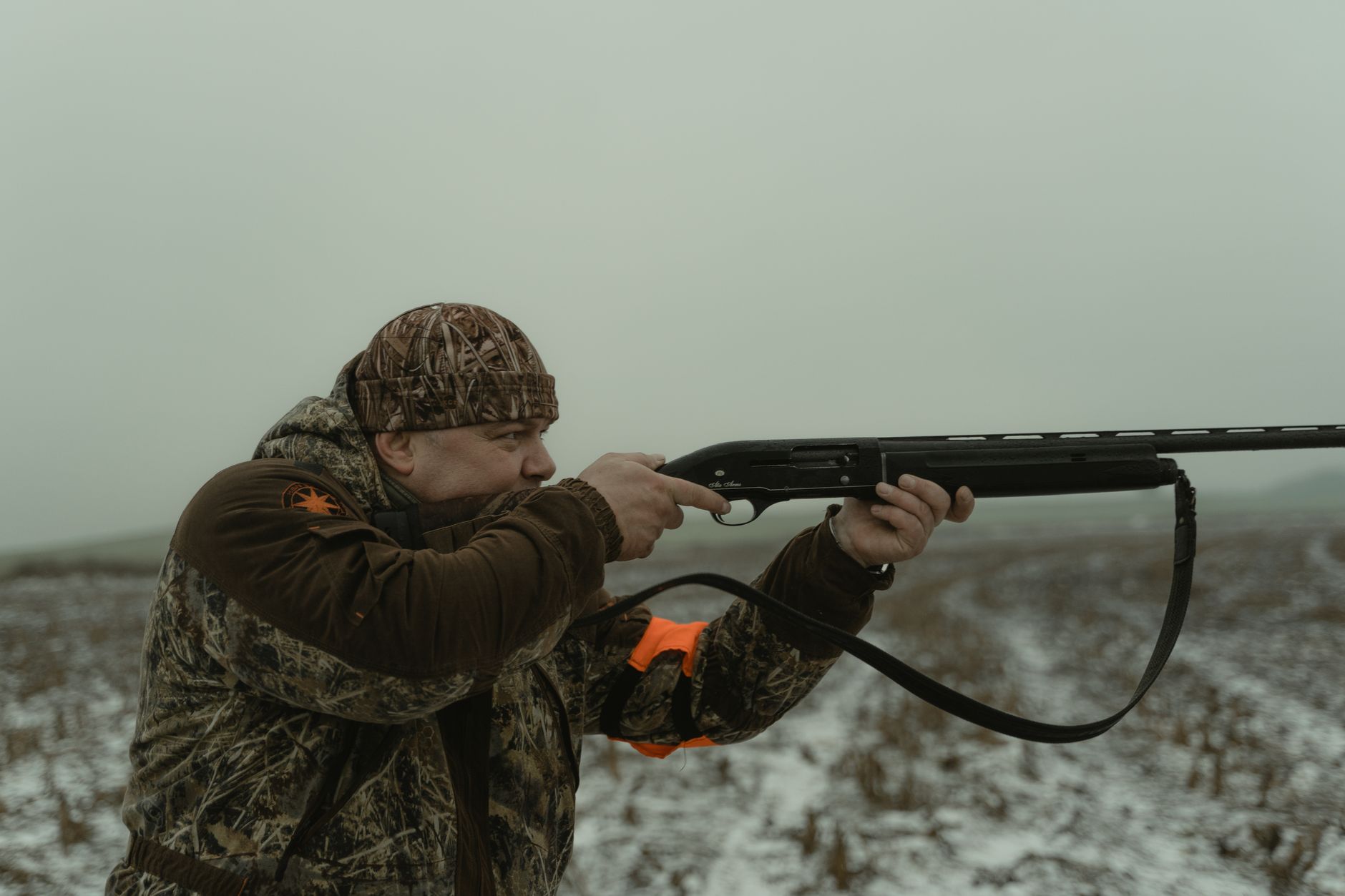 Hunter in camouflage gear aiming shotgun in a snowy field during winter hunting season.