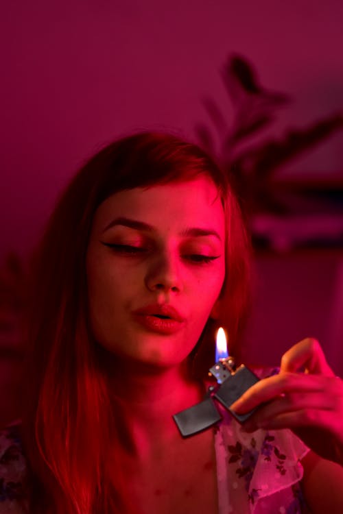 Serious female with long hair blowing out burning lighter with flame while sitting in obscure room with glowing red light at home