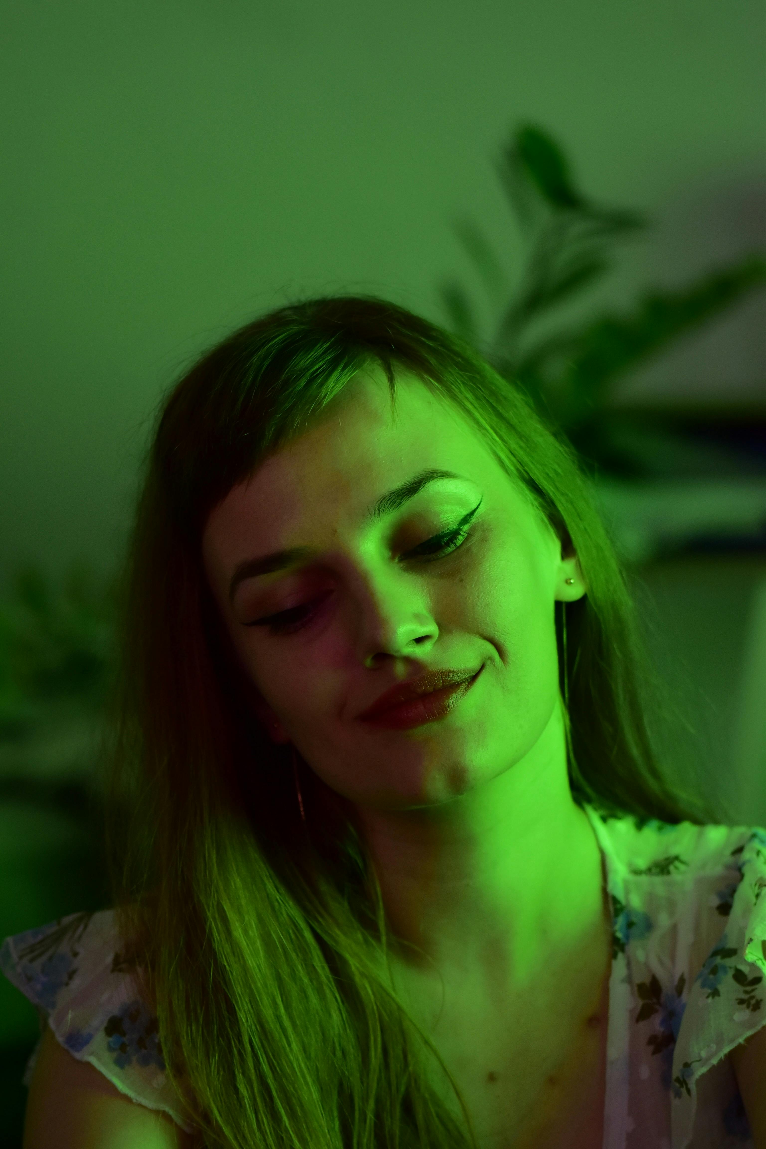 Charming young female with makeup smiling and looking down while sitting in room with plant and green light at home
