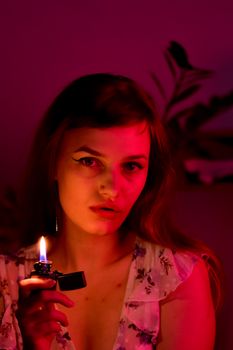 Calm female with long hair looking at camera while sitting with burning lighter in room with red illumination at home