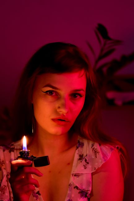 Calm female with long hair looking at camera while sitting with burning lighter in room with red illumination at home