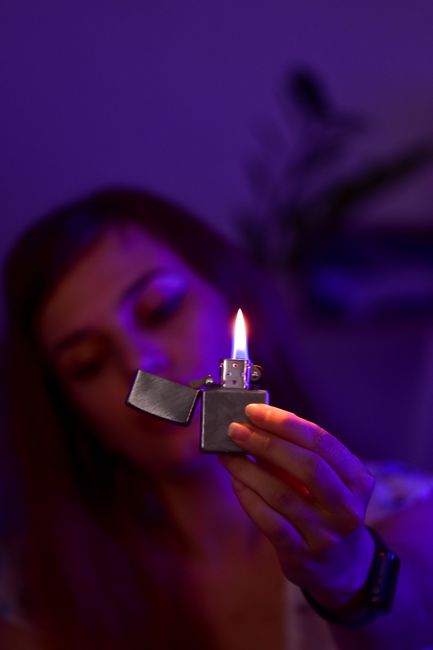 Blurred female with closed eyes demonstrating burning lighter in hand while sitting in dark room with purple illumination at home