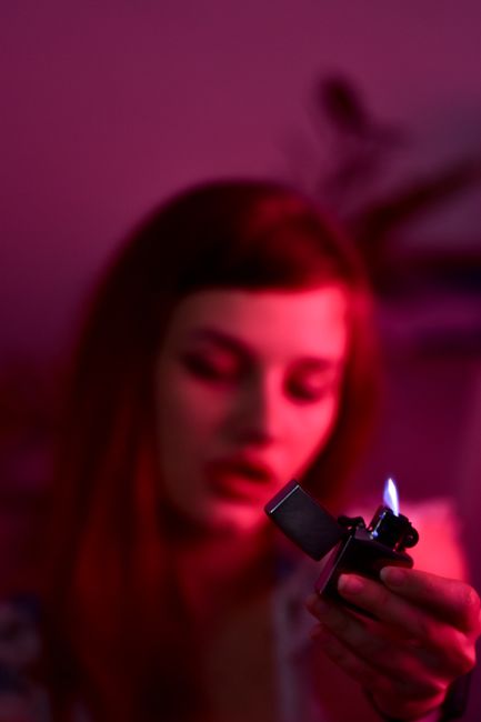 Blurred tranquil female with closed eyes and burning lighter with hot flame sitting in obscure room with red illumination at home