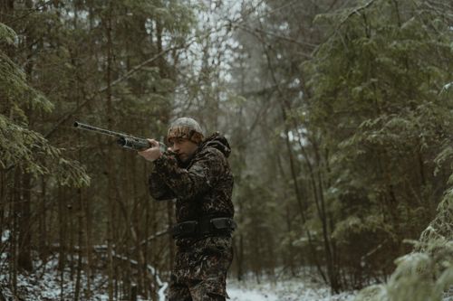 Free A man in camo gear aiming a rifle in a snowy forest, showcasing winter hunting. Stock Photo