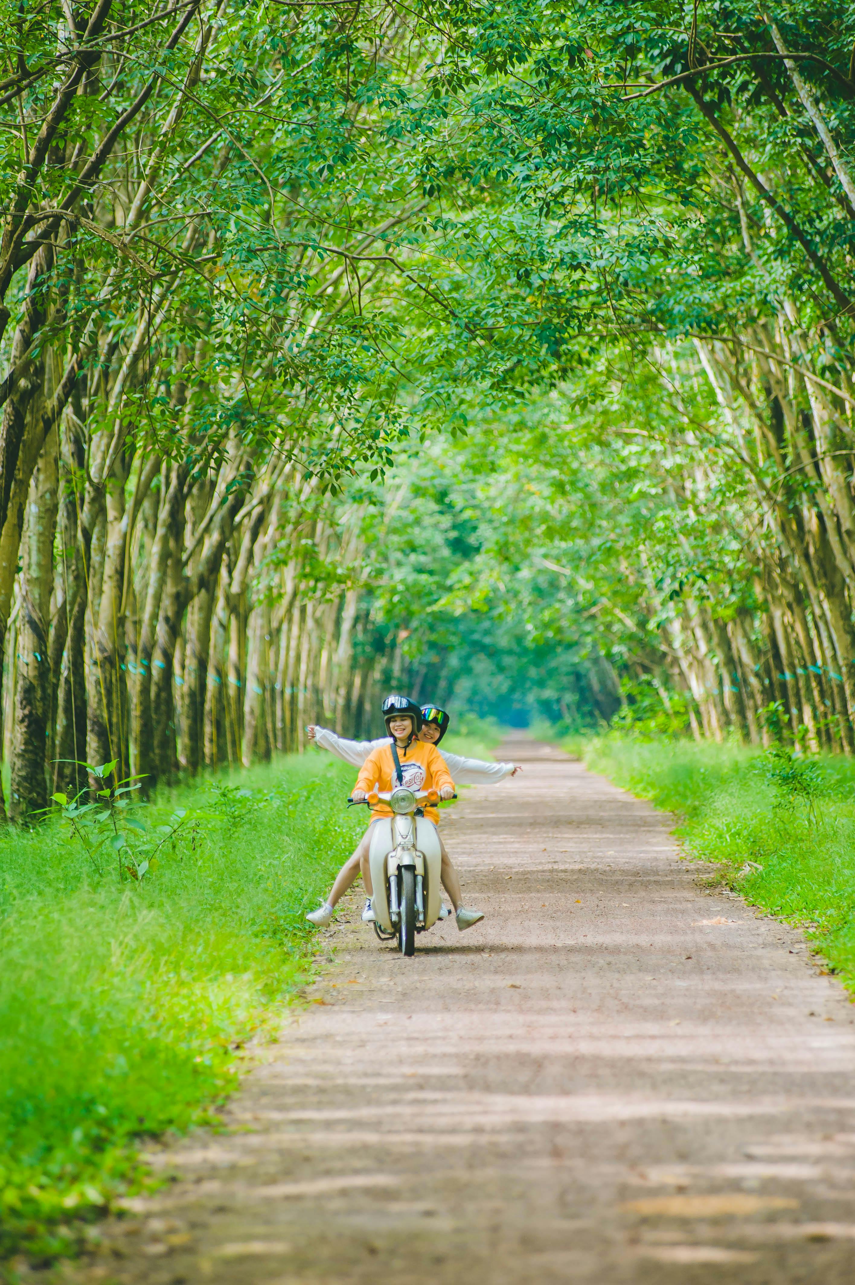 A couple joyfully riding a motorbike on a scenic, tree-lined dirt path.