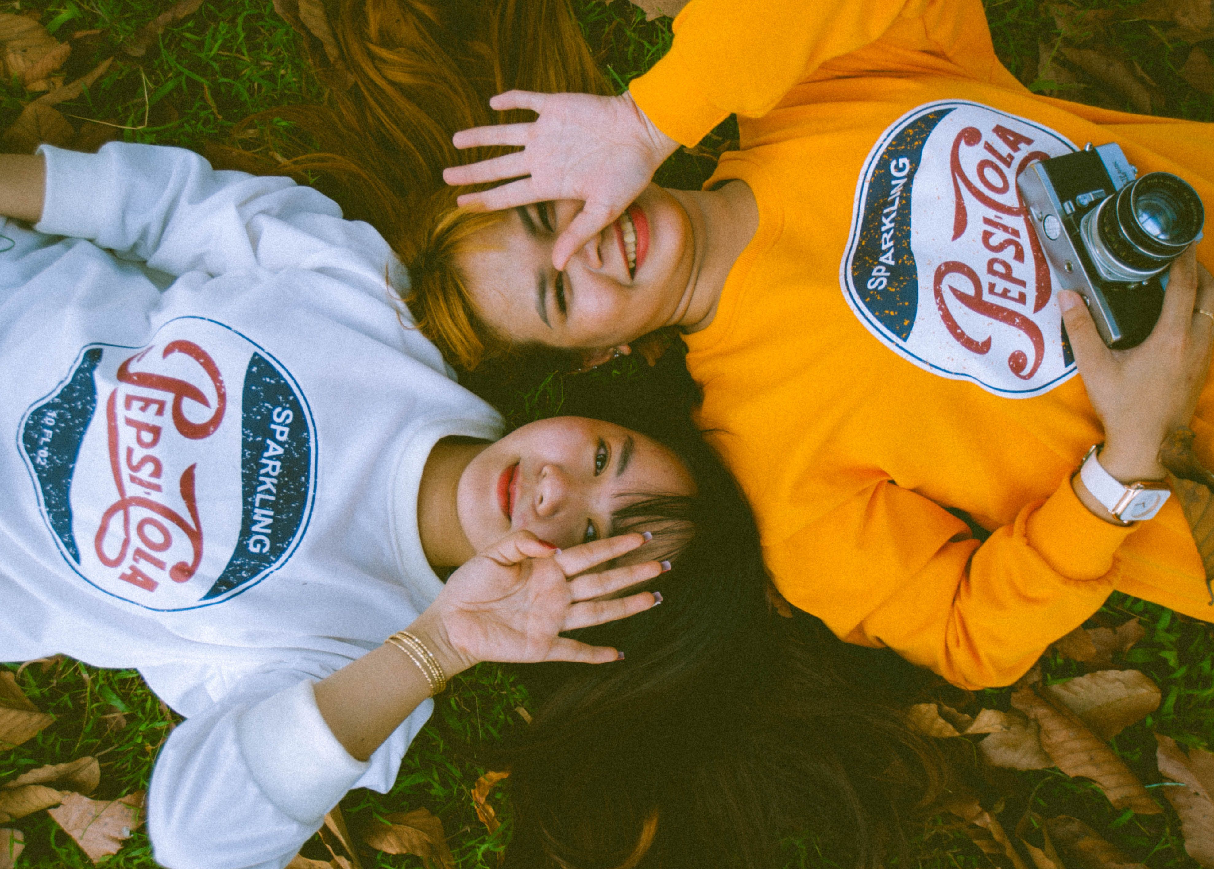 Two women lying on autumn leaves, smiling and holding a camera.