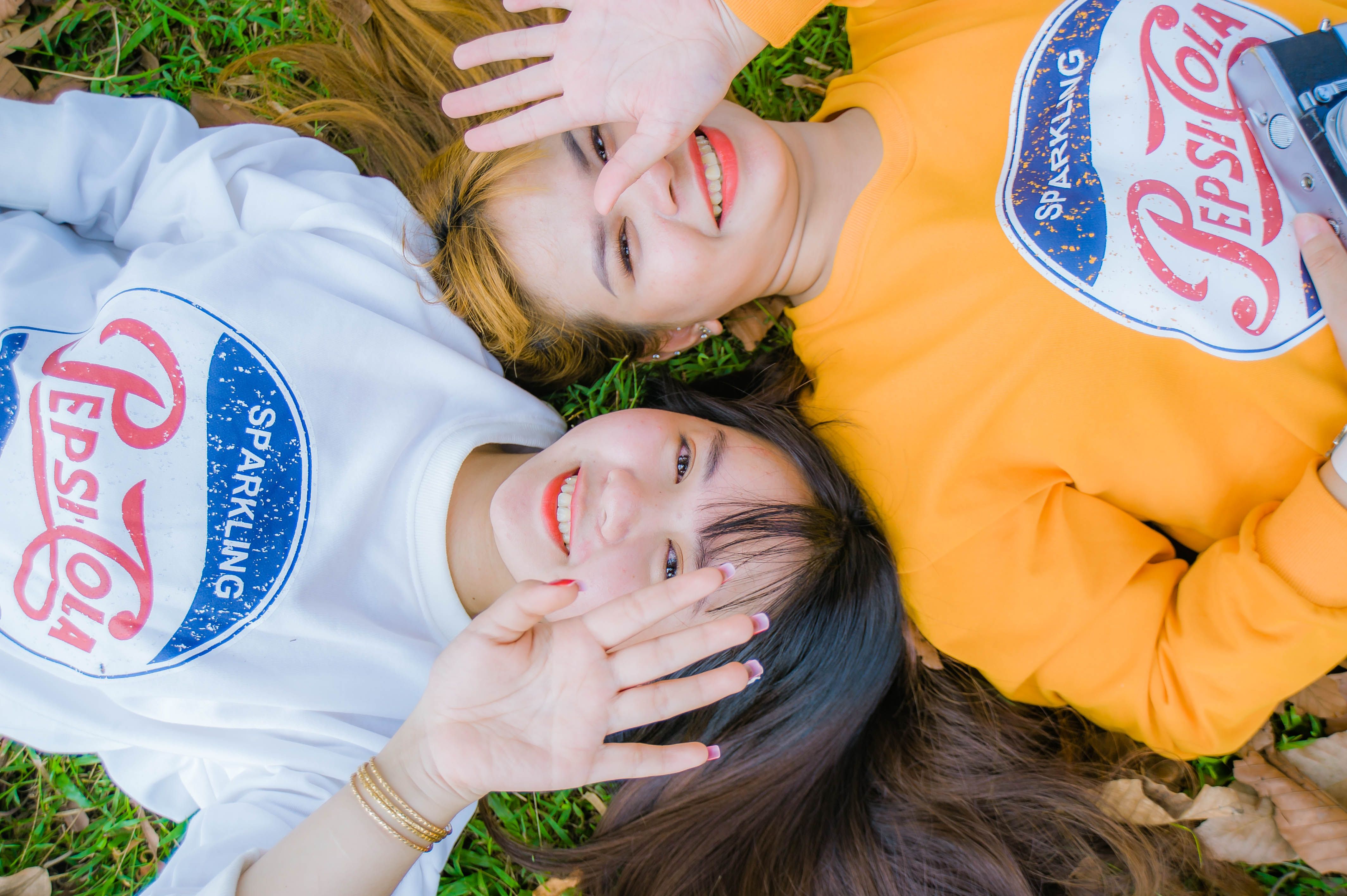 Two young women lying on grass, smiling playfully with Pepsi-Cola shirts.